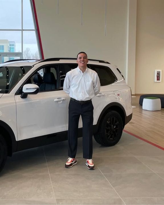 Mario in a white dress shirt beside a white SUV on a dealership showroom floor